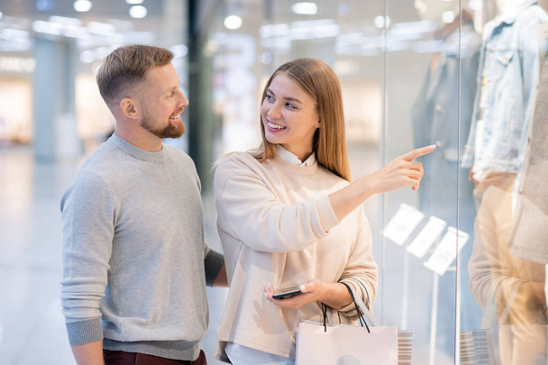 Młoda kobieta wskazująca płaszcz z nowej kolekcji sezonowej w witrynie sklepowej, patrząc z uśmiechem na męża podczas zakupów - Zdjęcie, obraz