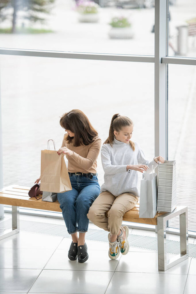 junge Frau und ihre Tochter sitzen auf Bank in großem Einkaufszentrum und betrachten Einkäufe in ihren Papiertaschen - Foto, Bild