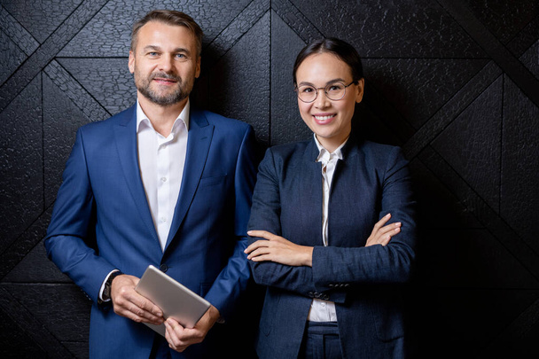 Happy successful colleagues in formalwear standing in front of camera by black wooden wall inside luxurious restaurant - Фото, изображение