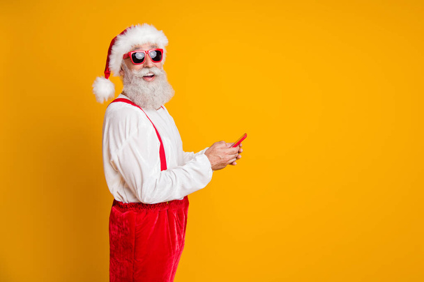 Profile side photo of funky white grey hair beard santa in red hat enjoy x-mas season noel discounts greeting he search online use smartphone isolated yellow color background - Photo, Image