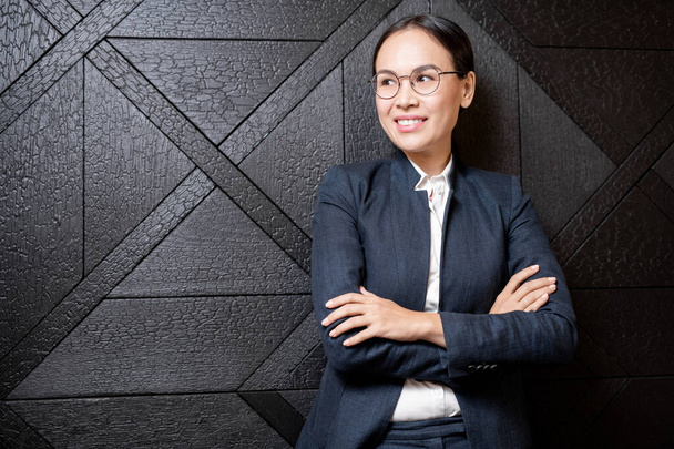 Young smiling Asian businesswoman with crossed arms standing by black wooden wall of luxurious restaurant - Фото, зображення