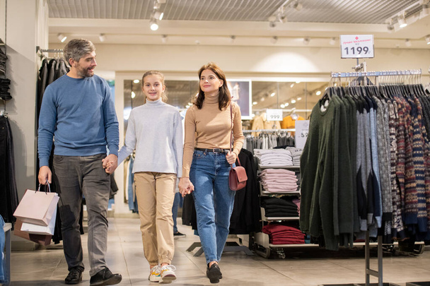 Famiglia felice di tre persone in tenuta casualwear a mano mentre si sposta lungo il reparto abbigliamento durante il periodo di vendita stagionale
 - Foto, immagini