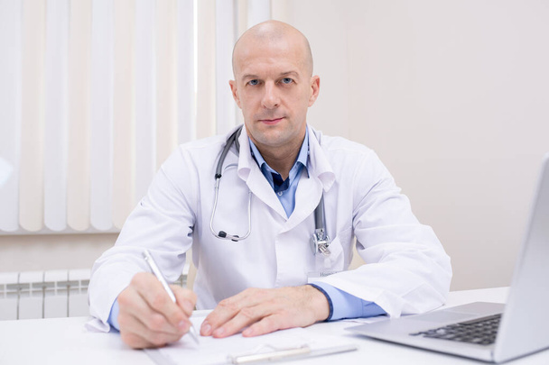 Médico confiante de meia idade careca em whitecoat sentado à mesa na frente da câmera e fazendo anotações no documento médico
 - Foto, Imagem