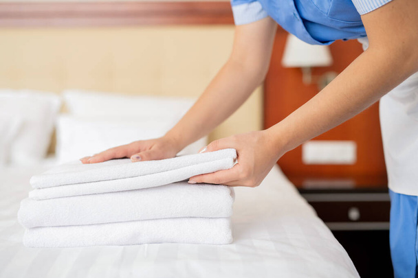 Genç oda hizmetçisinin elleri beyaz temiz havlu yığını yaparken yatağın üzerine eğilmiş. - Fotoğraf, Görsel