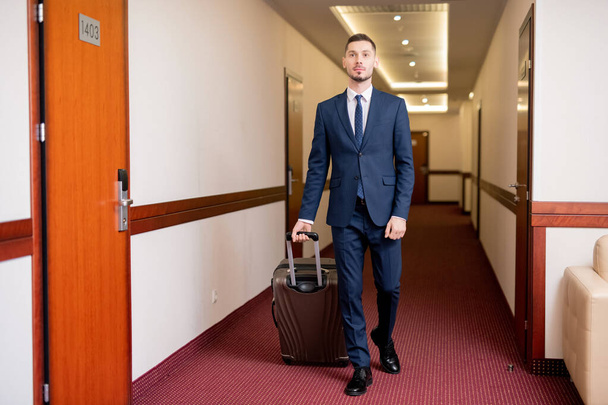 Současný dobře oblečený podnikatel tahá kufr se zavazadly a pohybuje se směrem ke svému pokoji v hotelu - Fotografie, Obrázek