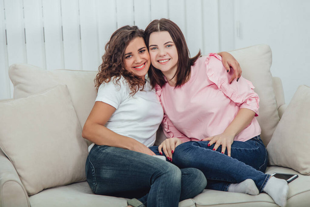Two amazing girls sitting on the sofa, hugging one another, smiling at the camera. - Fotó, kép