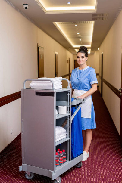 Красивая молодая горничная в форме стоит в длинном коридоре, получая чистые полотенца и другие вещи в гостиничных номерах
 - Фото, изображение