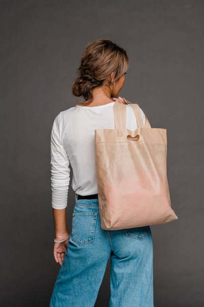Vista posterior de la joven hembra en ropa casual sosteniendo el bolso de mano beige desnudo en el hombro mientras está de pie en aislamiento
 - Foto, Imagen