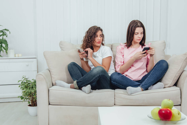 Two attractive girlfriends sitting on the sofa side by side in lotos position, with smartphones in hands. - Zdjęcie, obraz