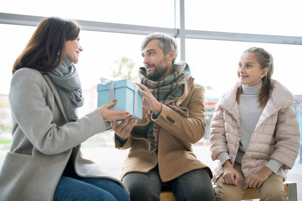 Σύζυγος σε casualwear δίνει τη σύζυγό του μπλε giftbox με χριστουγεννιάτικο δώρο με την κόρη τους κάθεται κοντά στο εμπορικό κέντρο - Φωτογραφία, εικόνα