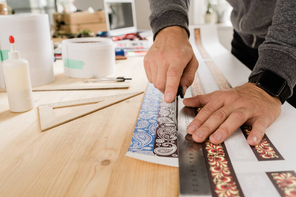 Χέρια του νέου τεχνίτη ή σχεδιαστή από ξύλινο τραπέζι χρησιμοποιώντας χάρακα και μαχαίρι για να κόψει διακοσμημένο χαρτί για να κάνει κολάρα - Φωτογραφία, εικόνα