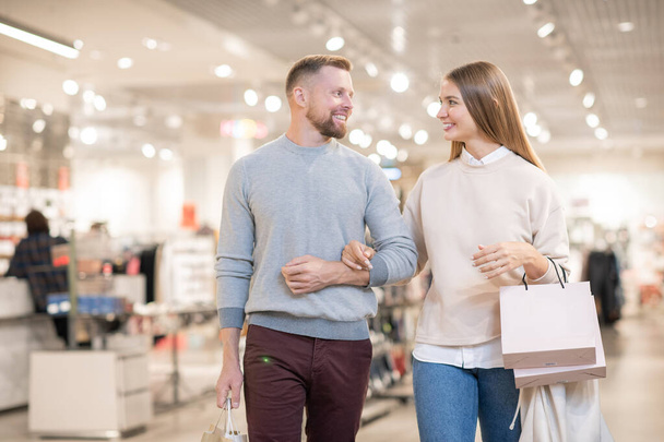 Alegre pareja mirándose mientras lleva bolsas de papel y dejando el departamento de ropa en el período de venta estacional
 - Foto, Imagen