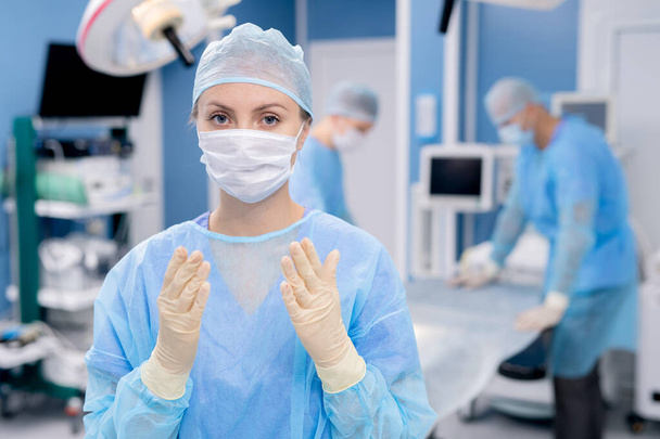 Νεαρός βοηθός με προστατευτική μάσκα, γάντια και στολή που σας κοιτάζει στο παρασκήνιο των συναδέλφων που ετοιμάζουν το χειρουργικό τραπέζι - Φωτογραφία, εικόνα