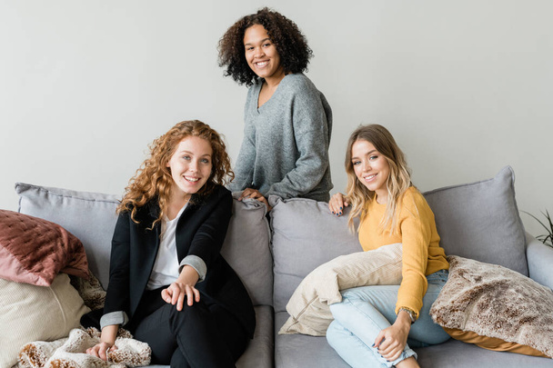 Group of cheerful friendly girls relaxing on soft comfortable sofa in front of camera and enjoying home rest - Photo, Image