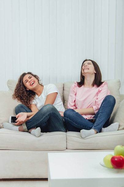 Két vonzó barátnő ül a kanapén lótusz pozícióban, okostelefonnal a kezükben, kétségbeesetten mosolyogva.. - Fotó, kép