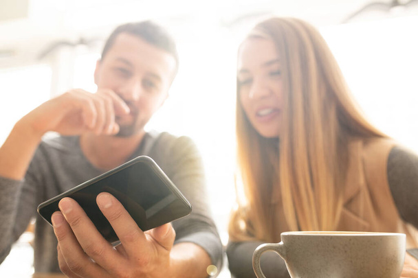 junger Mann hält Smartphone in der Hand, während er seiner Freundin oder Kollegin bei einer Tasse Tee im Café neugierige Sachen zeigt - Foto, Bild