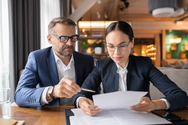 Confident businessman pointing at contract while his female colleague reading its terms and points at meeting in restaurant - 写真・画像