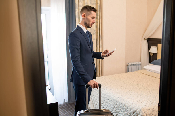 Młody, zajęty przedsiębiorca w stroju formalnym korzystający ze smartfona, aby zadzwonić po taksówkę, wyjeżdżając z hotelu na lotnisko - Zdjęcie, obraz
