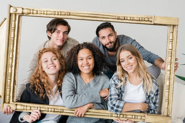 Группа молодых межкультурных веселых друзей в повседневной одежде смотрит на тебя, держа в руках большую портретную рамку
 - Фото, изображение