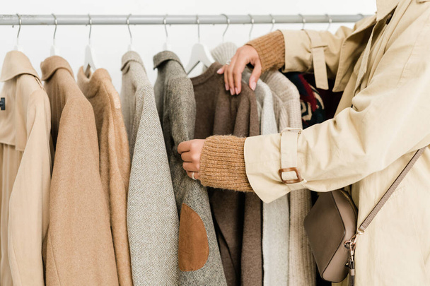Jeune femme contemporaine en trenchcoat beige et pull tricoté choisissant des vêtements décontractés de la nouvelle collection saisonnière en boutique
 - Photo, image