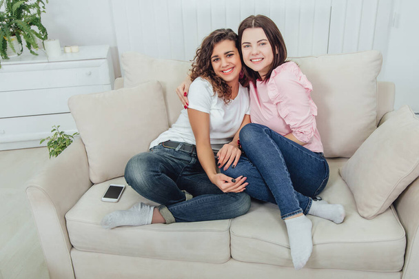 Two amazing girls sitting on the sofa, hugging one another, smiling lovely, looking at the camera. - Foto, Bild
