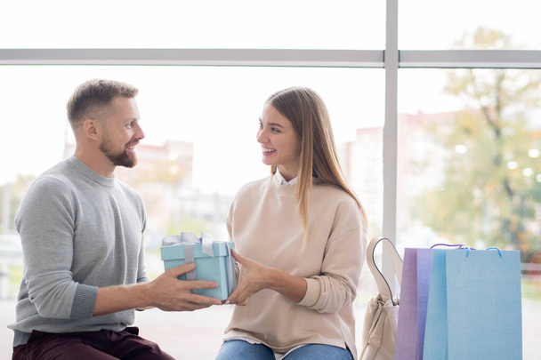 Усміхнена молода жінка бере подарункову коробку від своїх рук чоловіка, а обидва відпочивають після покупки біля вікна торгового центру
 - Фото, зображення