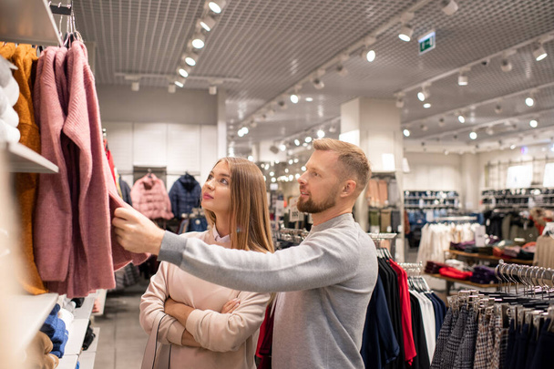 junges zeitgenössisches Paar steht neben einer neuen Casualwear-Kollektion auf Kleiderbügeln, während es sich einen von ihnen in der Bekleidungsabteilung ansieht - Foto, Bild