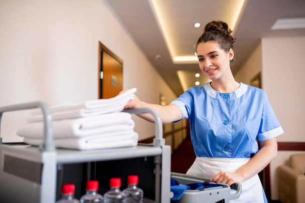 Young smiling chamber maid taking white clean towel from top of stack while pushing cart with hygiene stuff - 写真・画像