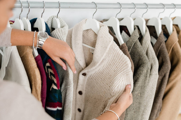 Manos de mujer joven sosteniendo cárdigan de punto blanco mientras elige ropa nueva en la boutique moderna
 - Foto, imagen