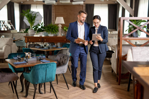 Dois parceiros de negócios elegantes caminhando pelo luxuoso restaurante e rolando no touchpad enquanto discutem dados on-line
 - Foto, Imagem