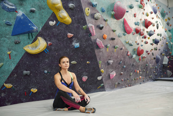 Mujer joven y serena con piernas cruzadas sentada en la alfombra en el gimnasio después de entrenar en el fondo del equipo de escalada
 - Foto, imagen