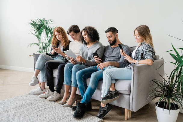 Row of contemporary casual guys and girls in casualwear using modern gadgets while relaxing on large softcouch - Photo, Image