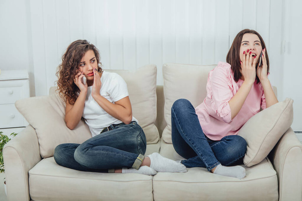 Two amazing girls sitting on the different sides of the sofa, talking on the phones. One girl is talking loudly and openly, another is keeping hand near mouth, speaking quietly. - Foto, imagen