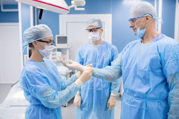 junge Krankenschwester hilft Professor in Schutzuniform, Handschuhe anzuziehen, während beide chirurgische Eingriffe vornehmen - Foto, Bild