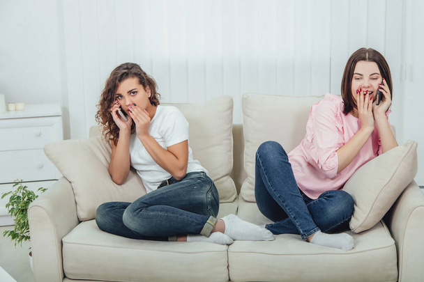 Two amazing girls sitting on the different sides of the sofa, talking on the phones. One girl is talking loudly and openly, another is keeping hand near mouth, speaking quietly. - Foto, Imagem