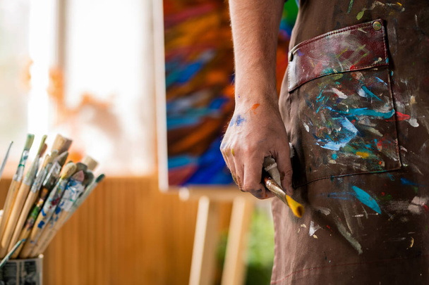 Hand van hedendaagse kunstenaar in schort met penseel terwijl hij werkt in zijn eigen atelier van kunst - Foto, afbeelding