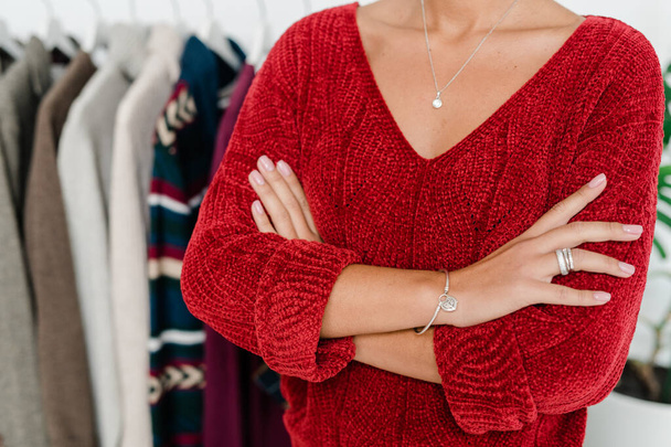 Молодая элегантная женщина в красном трикотаже пуловер скрещивая руки за грудь на фоне стойки с новой коллекцией одежды casualwear
 - Фото, изображение