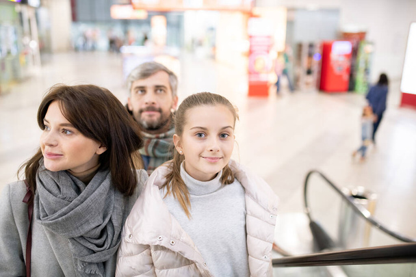 hübsches Mädchen und ihre Eltern bewegen sich auf der Rolltreppe nach oben, während sie während des saisonalen Verkaufs ein großes Handelszentrum besuchen - Foto, Bild