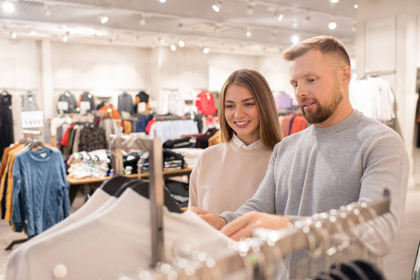 Веселая девушка и ее парень просматривают свитера или толстовки в отделе повседневной одежды в торговом центре или торговом центре
 - Фото, изображение