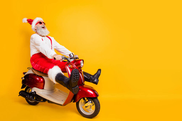 Full body photo of crazy santa white hair grandpa rushing x-mas party speed by retro bike wear trendy sun specs red trousers cap shirt boots isolated yellow color background - Zdjęcie, obraz