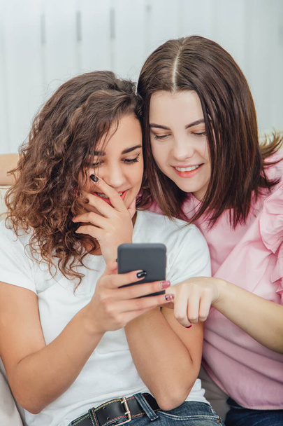 Beautiful young girls sitting on the sofa, at home, looking at the pictures in the phone, smiling, interacting. One girl is holding phone, another is pointing her finger at the screen. - Photo, image
