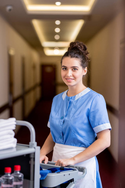Jovem sorridente morena feminina de uniforme azul olhando para você enquanto se move ao longo do corredor no hotel e empurrando carrinho com material de higiene
 - Foto, Imagem