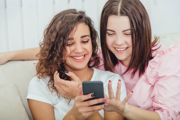 Beautiful young girls sitting on the sofa, at home, looking at the pictures in the phone, smiling, interacting. One girl is holding phone, another is pointing her finger at the screen. - Фото, изображение