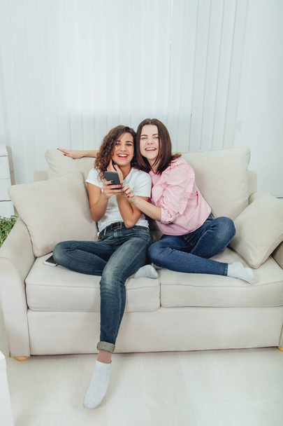 Beautiful young girls sitting on the sofa, at home, looking at the pictures in the phone, smiling, looking at the camera. One girl is holding phone, another is pointing her finger at the screen. - Фото, зображення