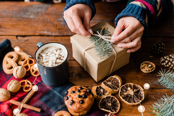 Hands of girl making knot on top of wrapped giftbox surrounded by sweet food, hot drink in mug and walnuts - Photo, Image