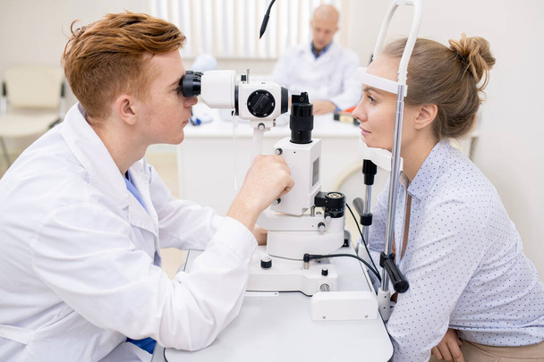 Młody okulista i jego pacjent siedzący przed sobą podczas badań okulistycznych sprzętu optycznego - Zdjęcie, obraz