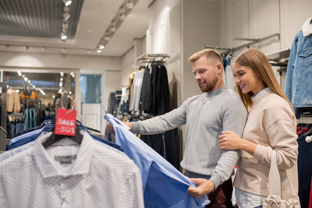 Jovem mulher e seu marido olhando para camisa azul, enquanto em pé por rack com etiqueta de desconto e escolher novas roupas
 - Foto, Imagem