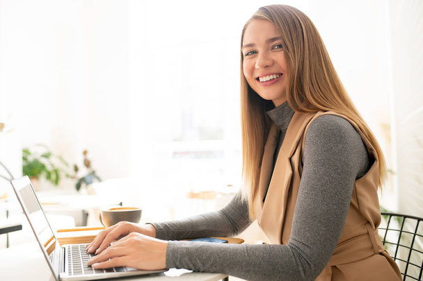 Молодая элегантная деловая женщина, глядя на тебя с зубастой улыбкой, печатая на клавиатуре ноутбука в кафе
 - Фото, изображение