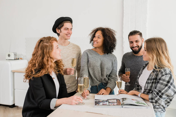 Grupo de caras e meninas amigáveis com flautas de champanhe conversando e olhando através da revista na festa em casa
 - Foto, Imagem