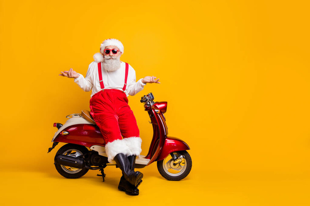 Full size photo of santa sit moped shrug shoulders not feel guilty coming party without gift wear specs suspenders pants cap shirt boots isolated yellow color background - 写真・画像
